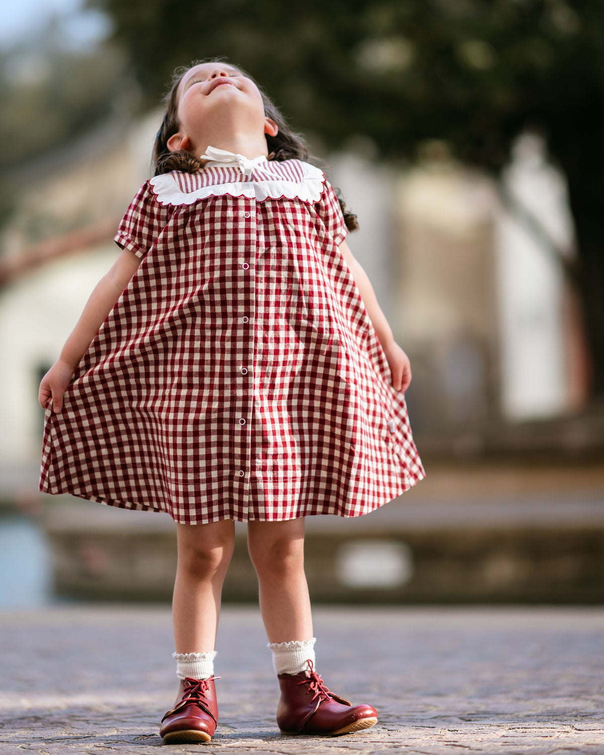 Young girl in a red and white checkered dress standing outdoors with trees in the background.