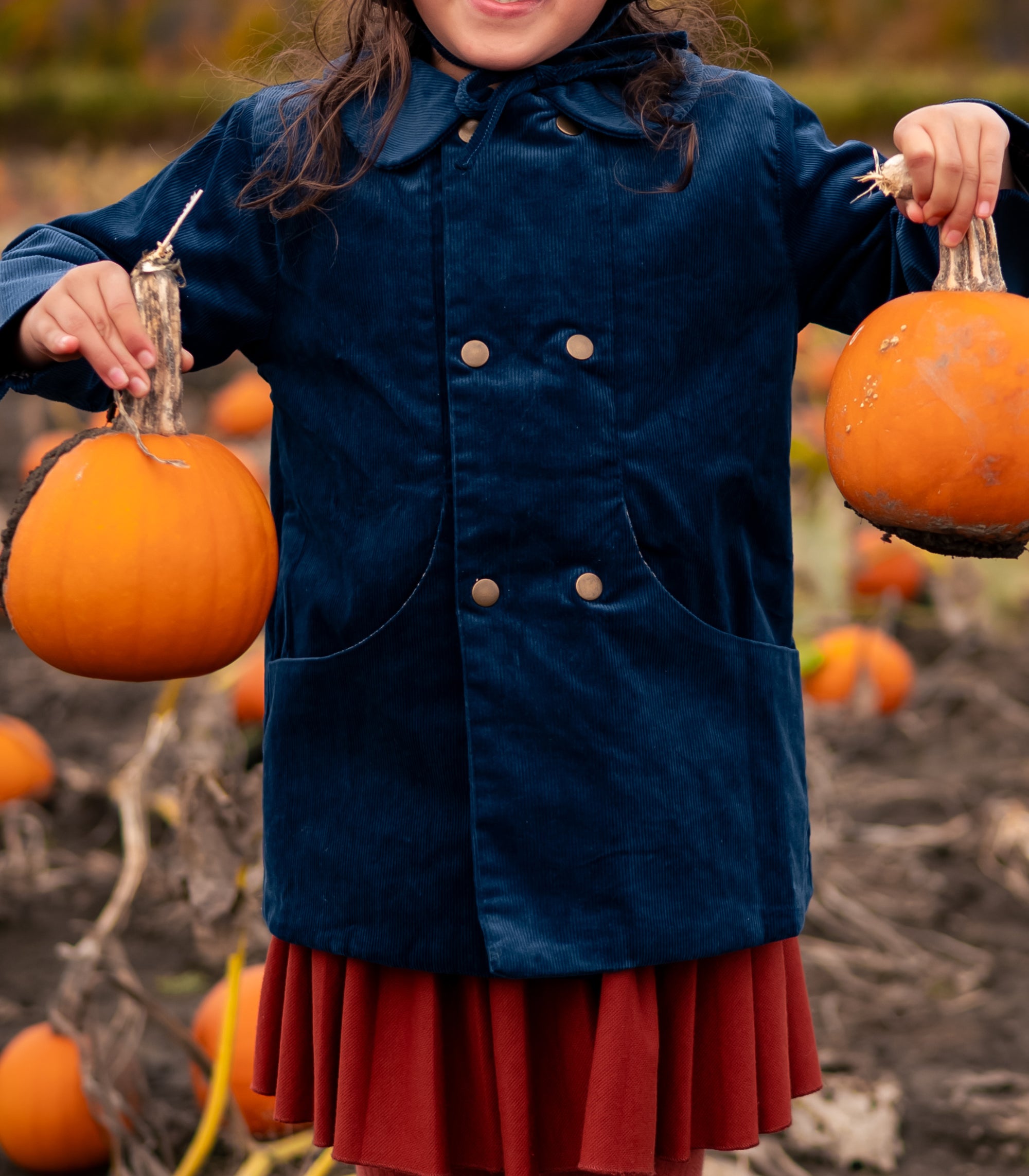 Girl holding two pumpkins in a pumpkin patch wearing a blue corduroy coat