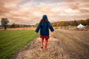 Girl in a blue coat and matching hat on hay bales in an open field with a cloudy sky.