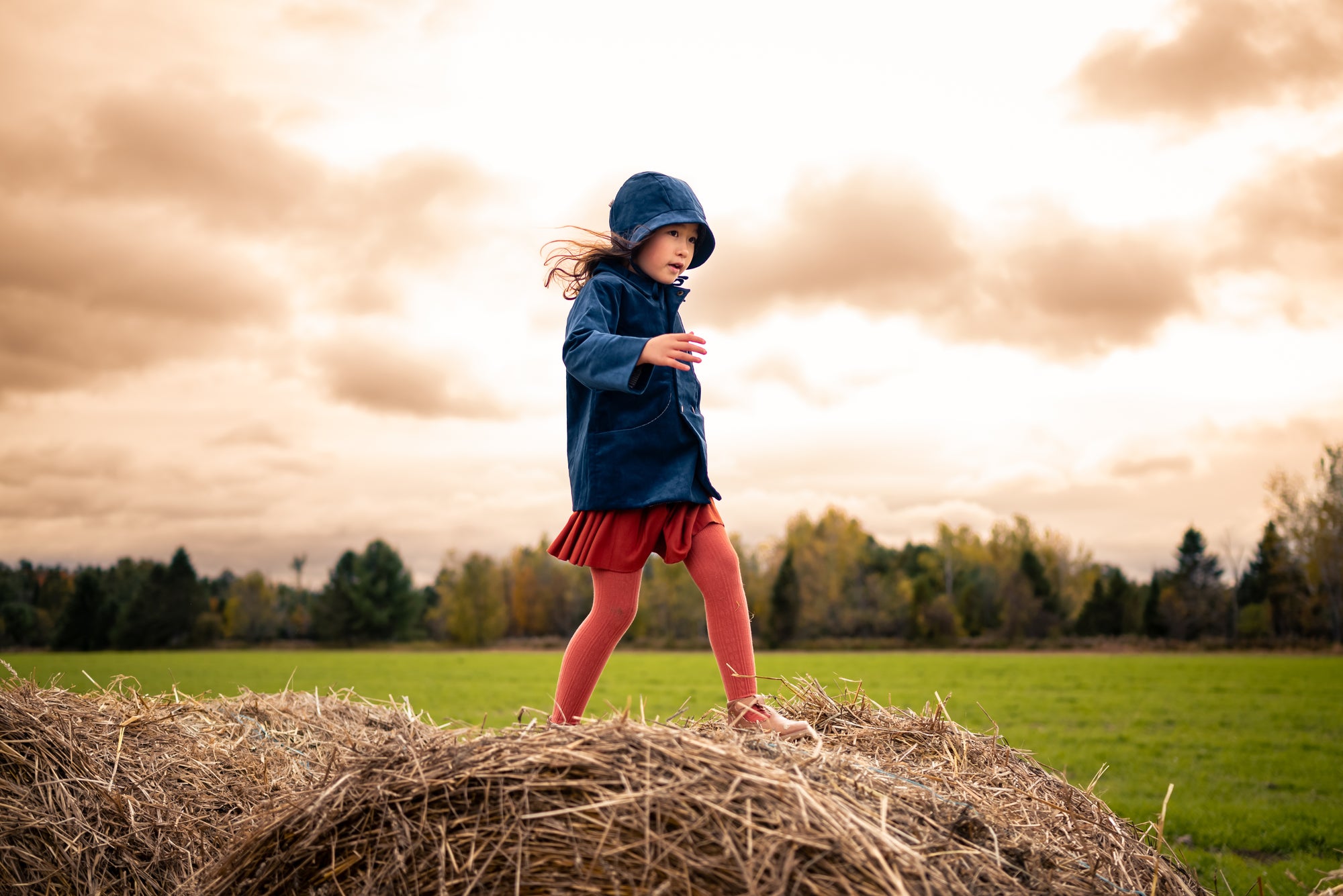 Child in a blue coat and red skirt standing on a hay bale with a cloudy sky and trees in the background.