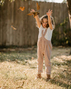 A young child wearing a white short-sleeve shirt and brown-and-white gingham check overalls stands outdoors in a backyard, throwing autumn leaves in the air. The background features a wooden fence, grass, and trees.