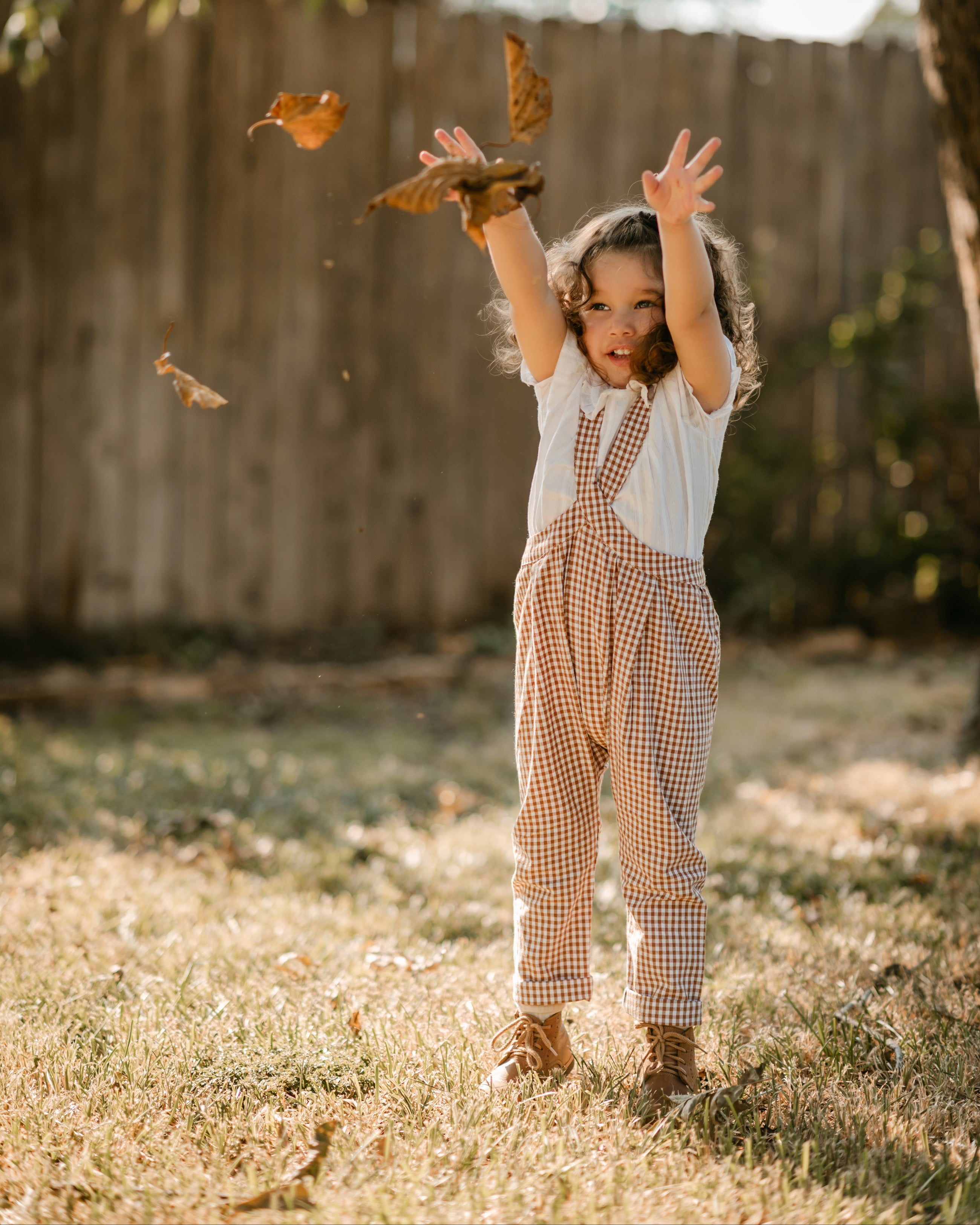 A young child wearing a white short-sleeve shirt and brown-and-white gingham check overalls stands outdoors in a backyard, throwing autumn leaves in the air. The background features a wooden fence, grass, and trees.
