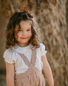A young girl with curly brown hair wearing a white ruffled blouse and brown gingham check overalls, standing outdoors in front of a textured tree trunk.
