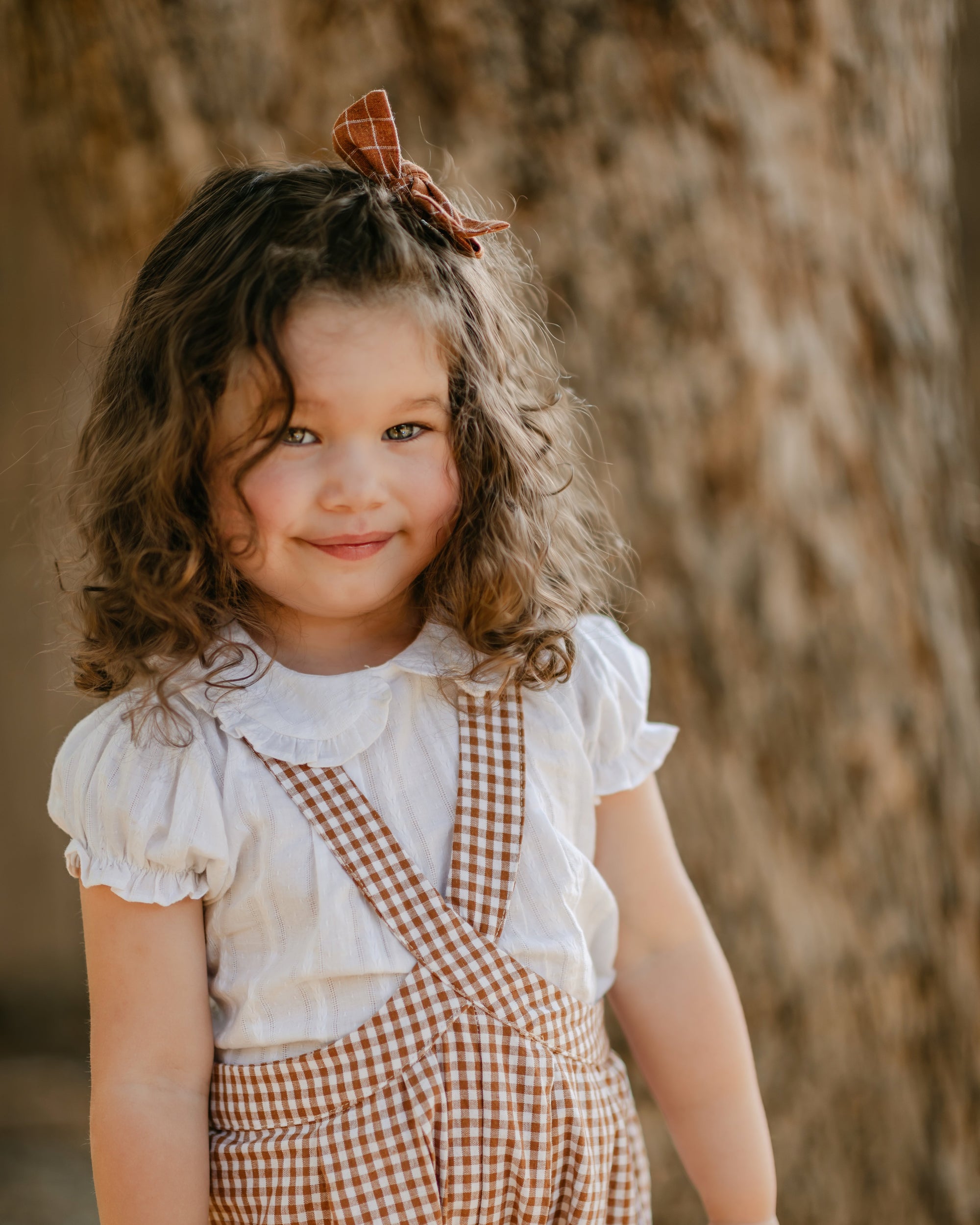 A young girl with curly brown hair wearing a white ruffled blouse and brown gingham check overalls, standing outdoors in front of a textured tree trunk.