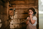 A young child in a white blouse and brown gingham pinafore dress stands beside a vintage wooden wall-mounted telephone, holding the receiver to their mouth in a rustic interior.