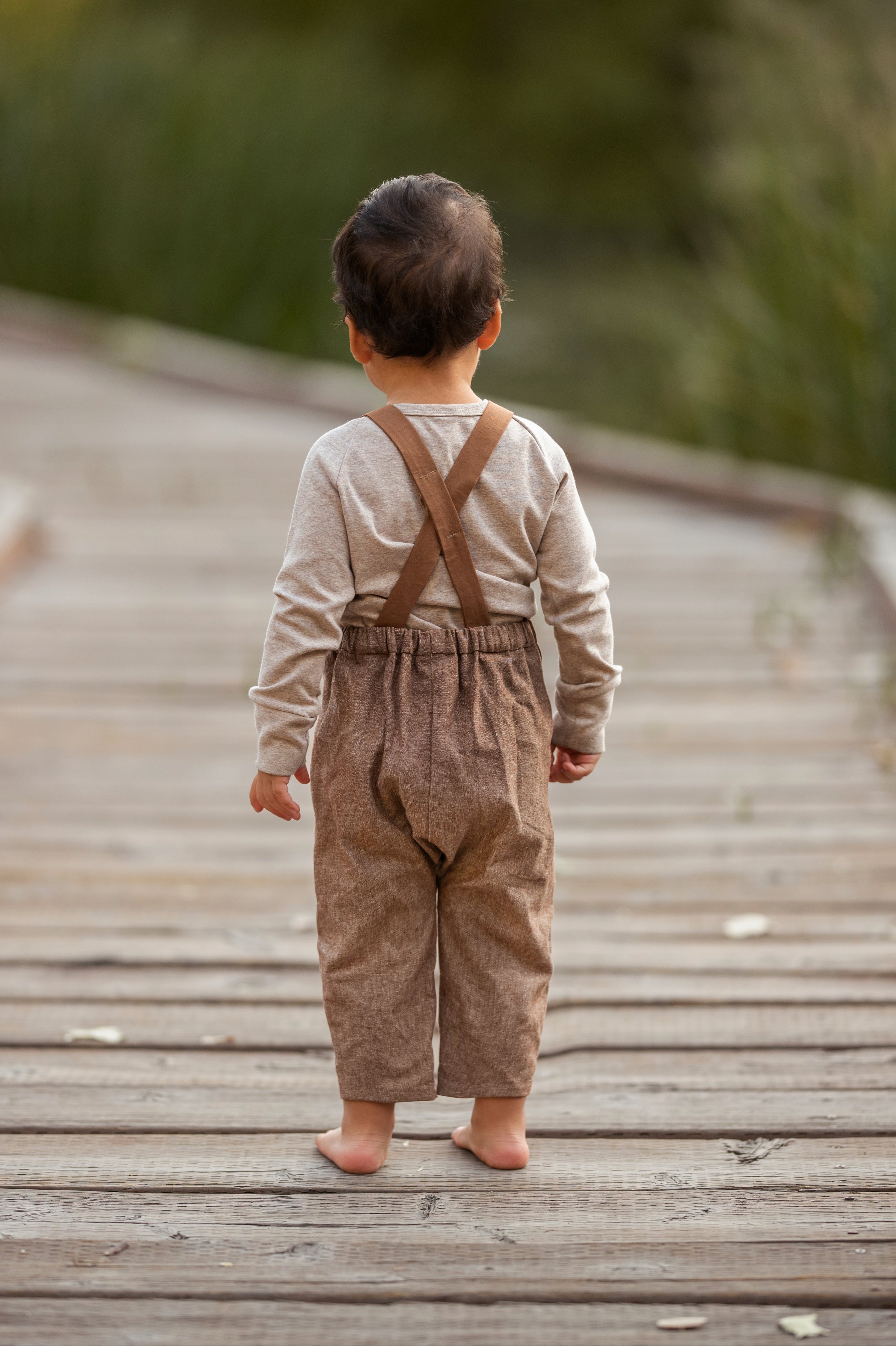 Child wearing brown suspenders and pants on a wooden pathway