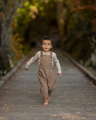 Child wearing brown overalls walking on a wooden path with a blurred natural background
