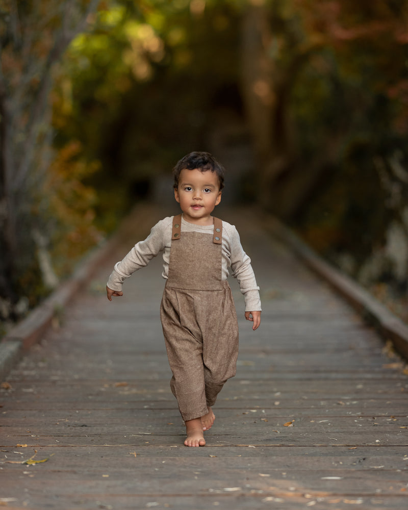 Child wearing brown overalls walking on a wooden path with a blurred natural background