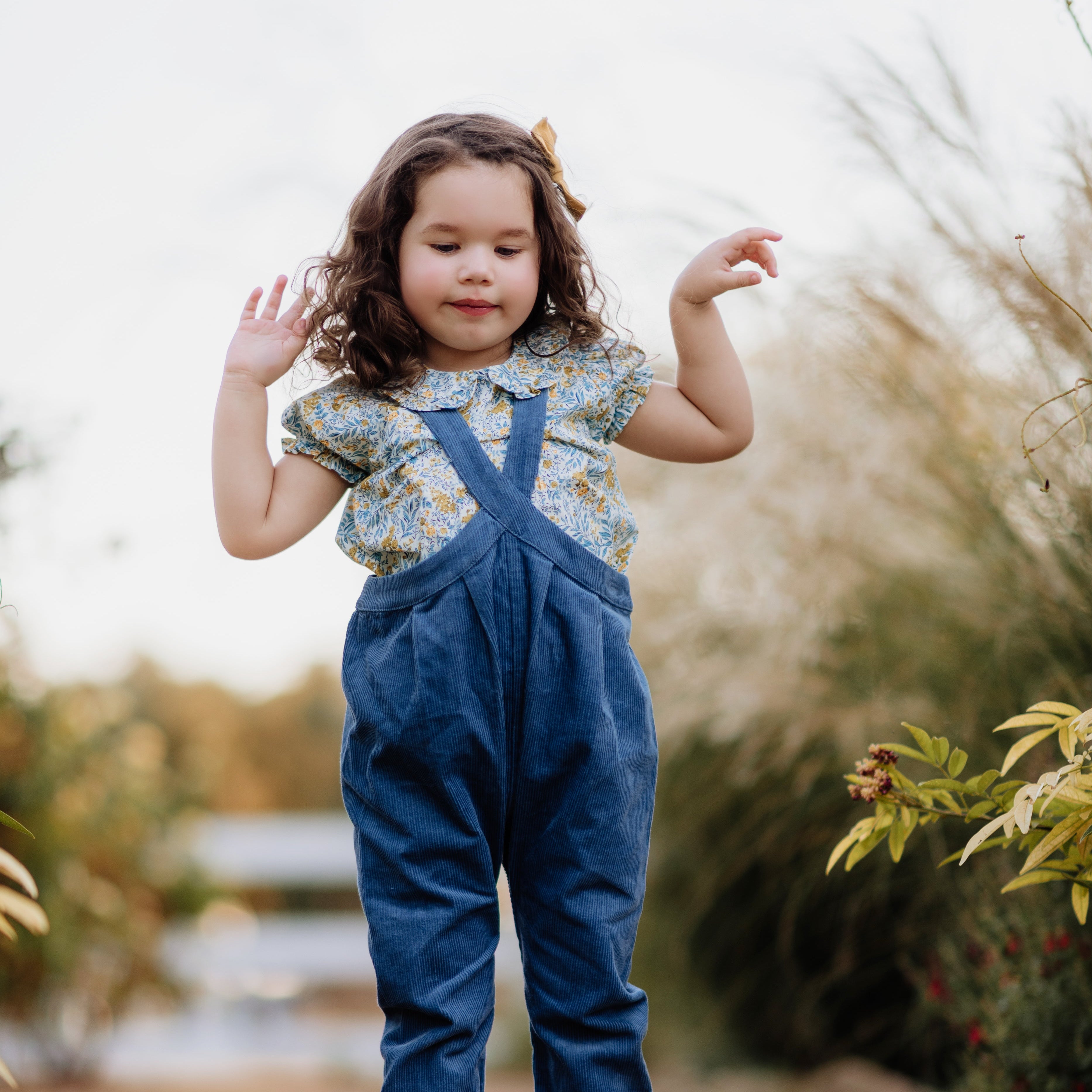 Young girl in blue overalls standing outdoors with a blurred natural background