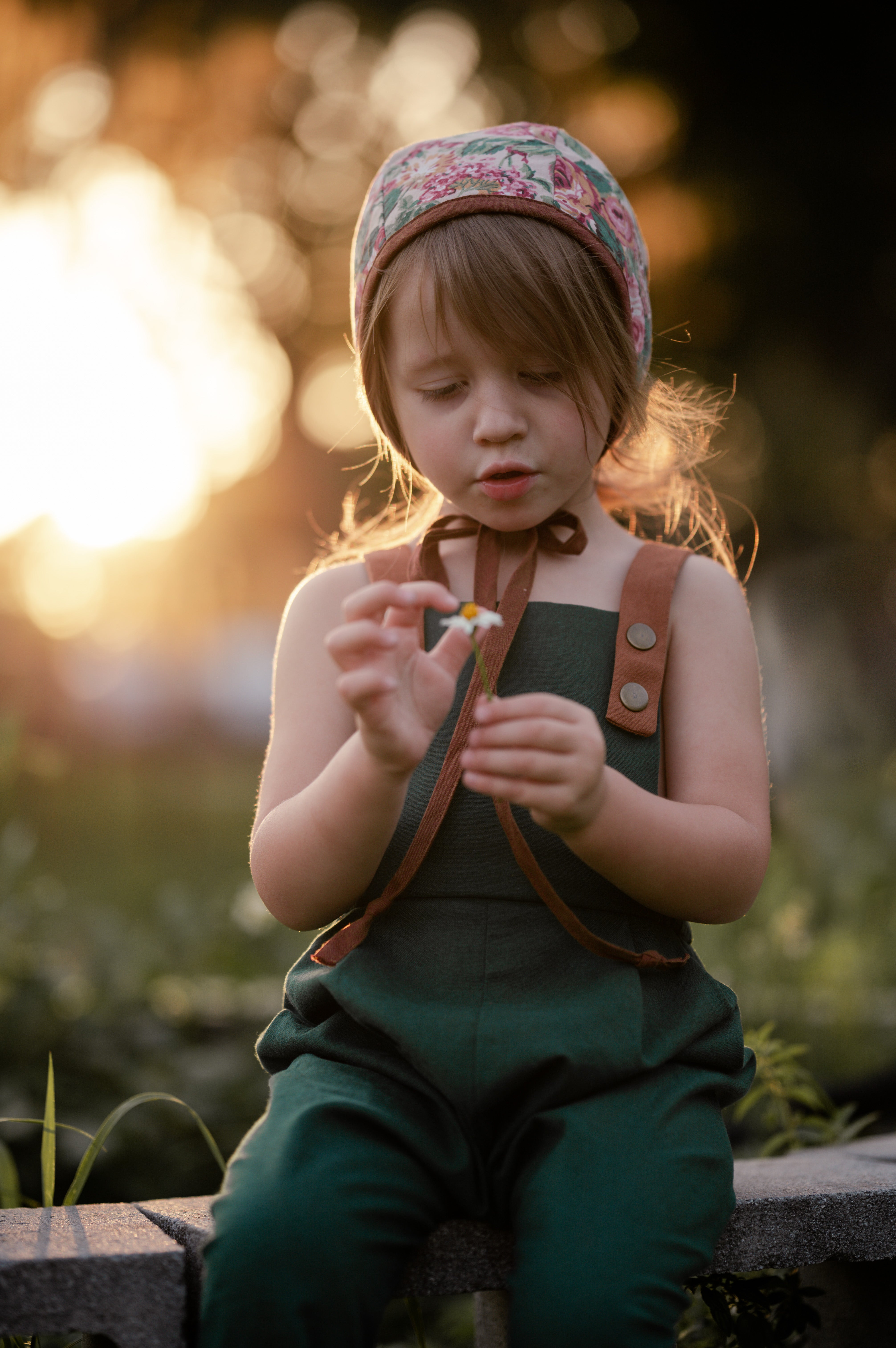 A 3-year-old girl sitting down, wearing the evergreen long overalls and a cute bonnet, playfully holding a daisy, highlighting comfort and charm in children’s fashion.