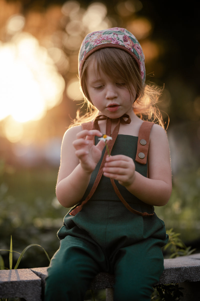 A 3-year-old girl sitting down, wearing the evergreen long overalls and a cute bonnet, playfully holding a daisy, highlighting comfort and charm in children’s fashion.