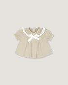 A baby girl's short-sleeve top in beige and white vertical stripes, featuring a white gingham Peter Pan collar, a white bow at the neckline, and a small white side tag, shown on a solid light gray background.