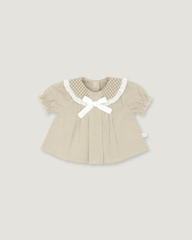 A baby girl's short-sleeve top in beige and white vertical stripes, featuring a white gingham Peter Pan collar, a white bow at the neckline, and a small white side tag, shown on a solid light gray background.