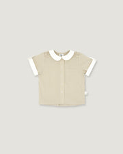 A short-sleeve baby shirt in beige ribbed fabric with a white Peter Pan collar, white sleeve cuffs, and a subtle checkered pattern on the shoulders, shown on a solid light gray background.