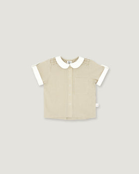 A short-sleeve baby shirt in beige ribbed fabric with a white Peter Pan collar, white sleeve cuffs, and a subtle checkered pattern on the shoulders, shown on a solid light gray background.