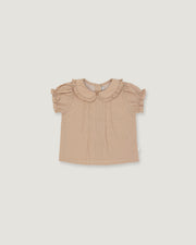A toddler girl's short-sleeve blouse in a light beige or dusty tan color, featuring a ruffled Peter Pan collar and ruffled sleeves, laid flat against a solid light gray background.