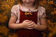 Child wearing a red apron with yellow flowers, standing against a blurred natural background.