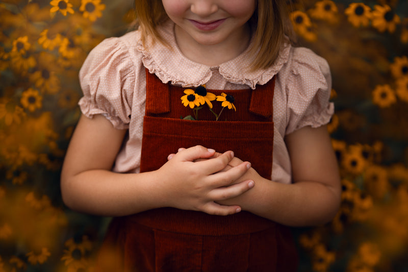 Child wearing a red apron with yellow flowers, standing against a blurred natural background.