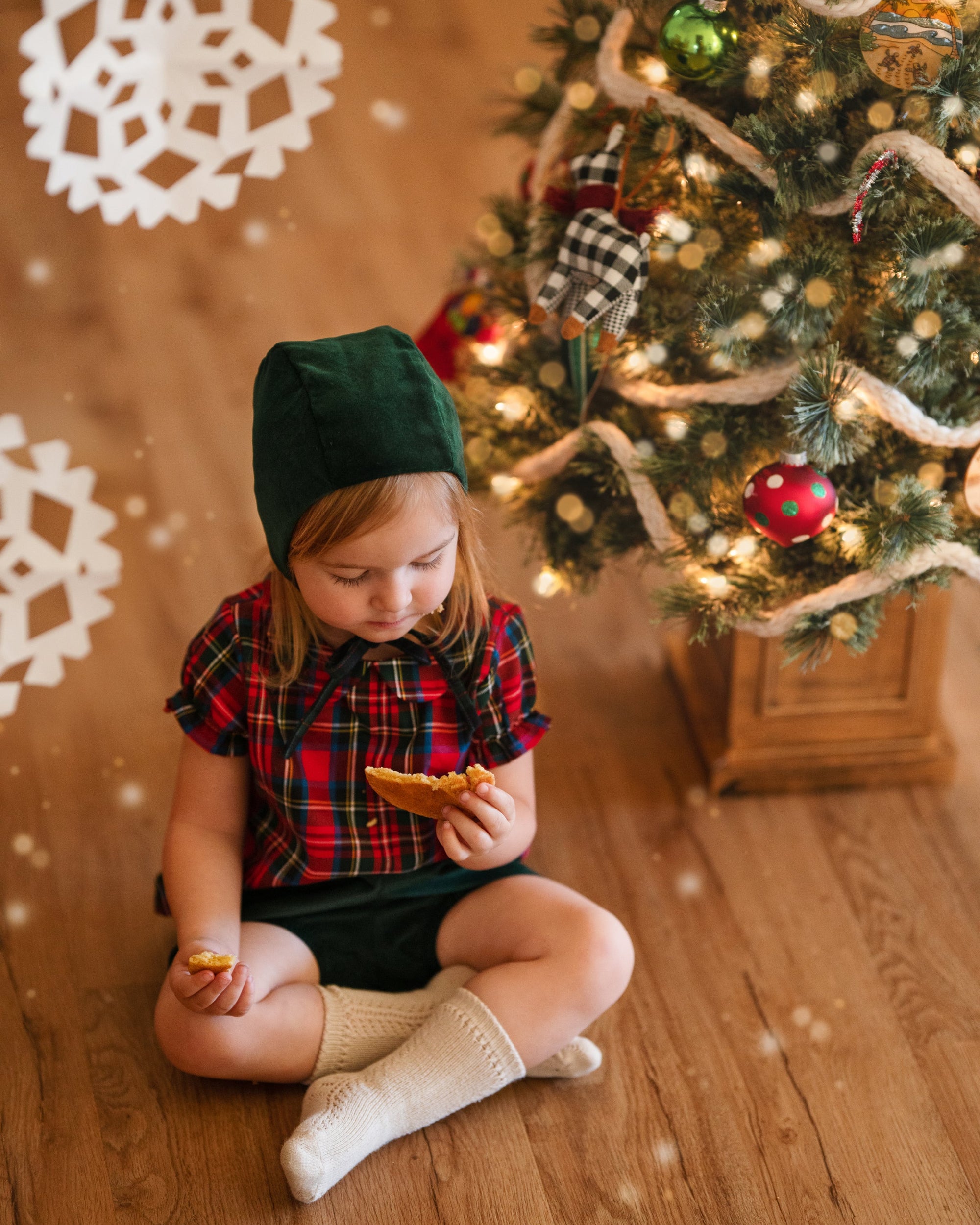 Girl in green bonnet and plaid blouse eating cooking in front of a Xmas tree