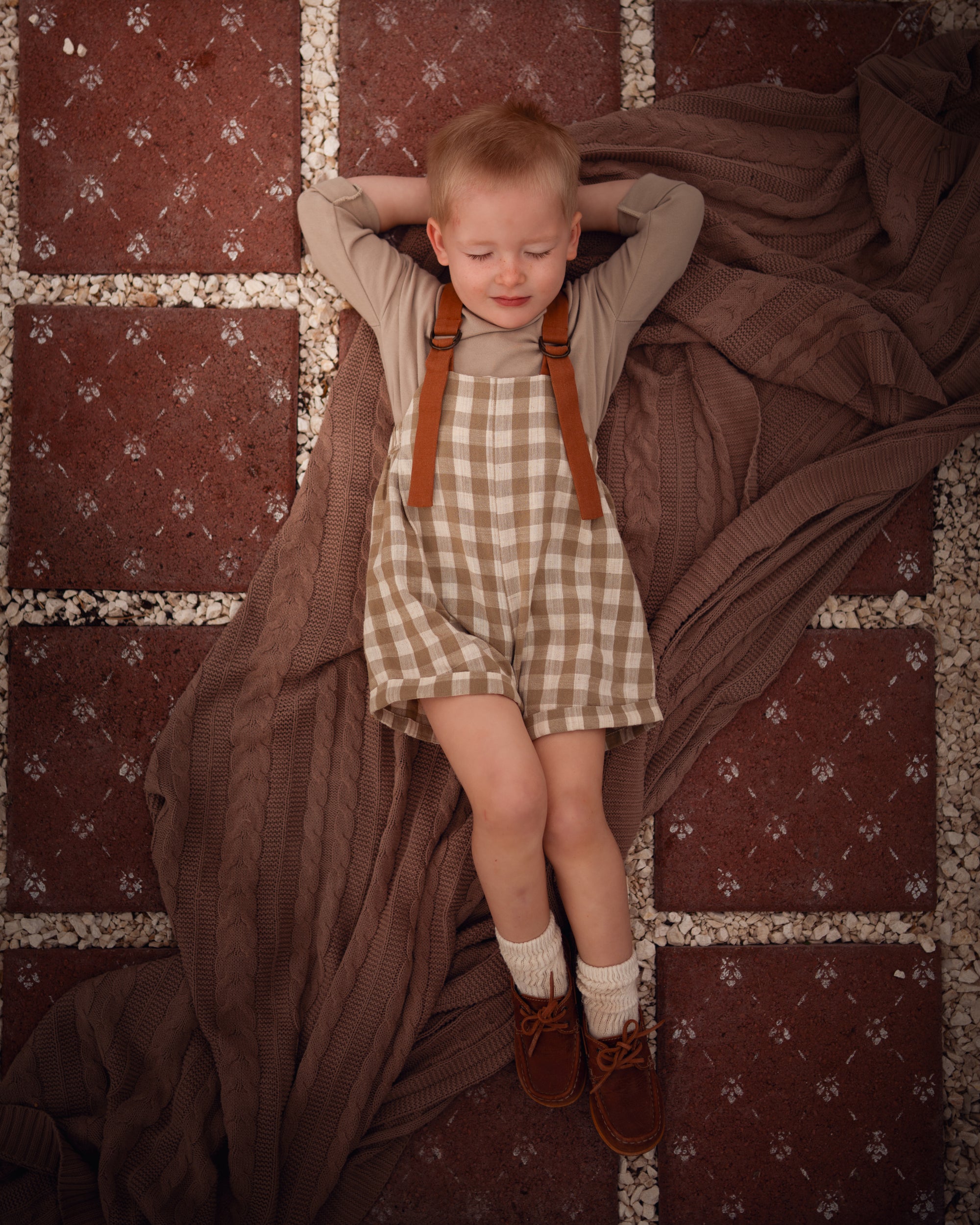 Model boy lying down in a Grow-With-Me Romper in Beige Gingham over a neutral top, showcasing the adjustable straps and classic gingham pattern, exuding a relaxed and playful vibe while emphasizing the romper's versatility and comfort.