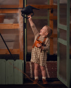 Model boy playing with a watering can in front of a green door, wearing the Grow-With-Me Romper in Beige Gingham over a neutral top, laughing joyfully as he enjoys a playful moment, highlighting the romper's comfort and style in a cheerful outdoor setting.
