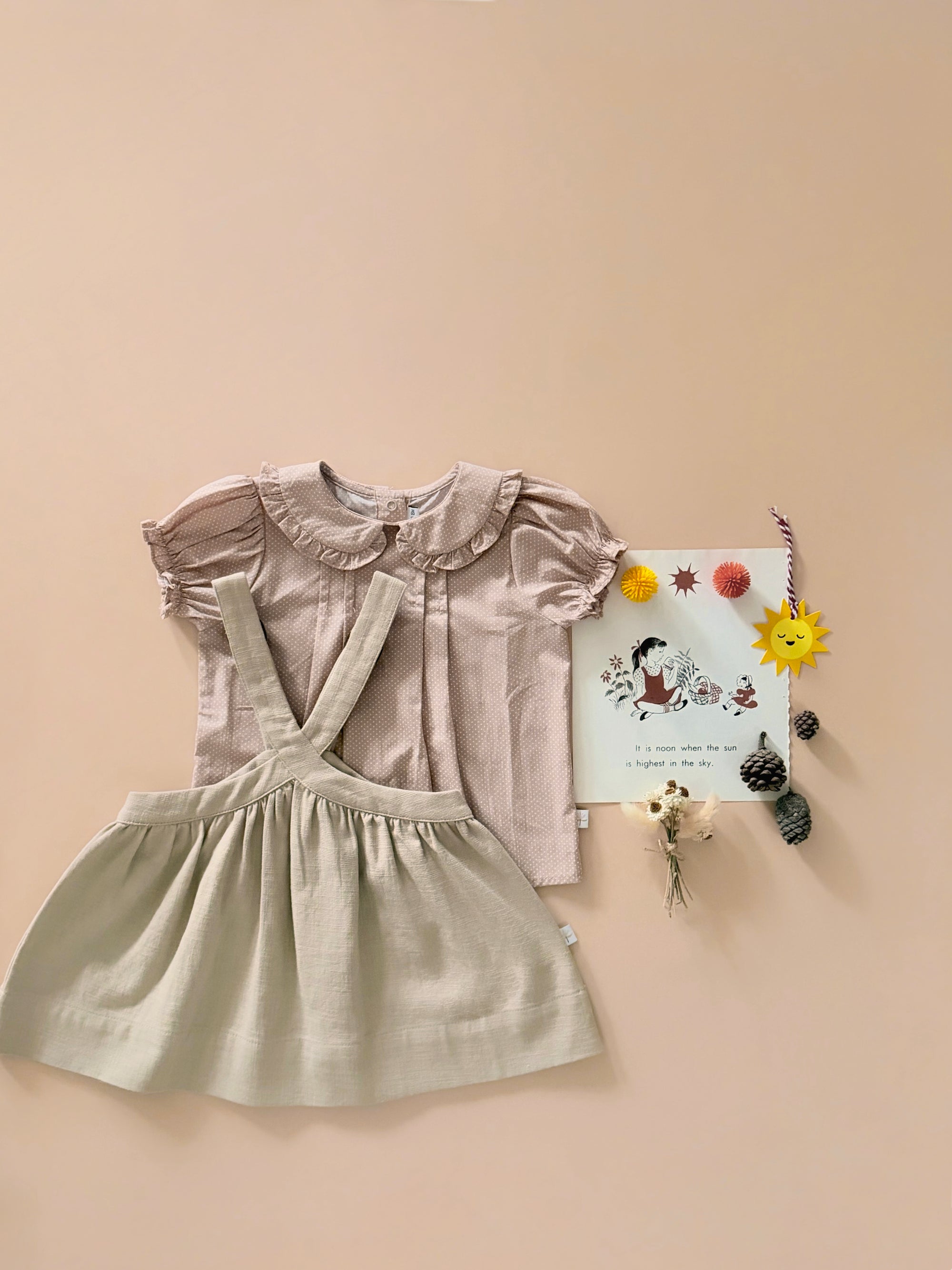 A two-piece children's outfit set featuring a dusty rose short-sleeve top with a ruffled collar and a light sage green linen-look overall skirt, styled flat-lay on a warm beige background with decorative props.