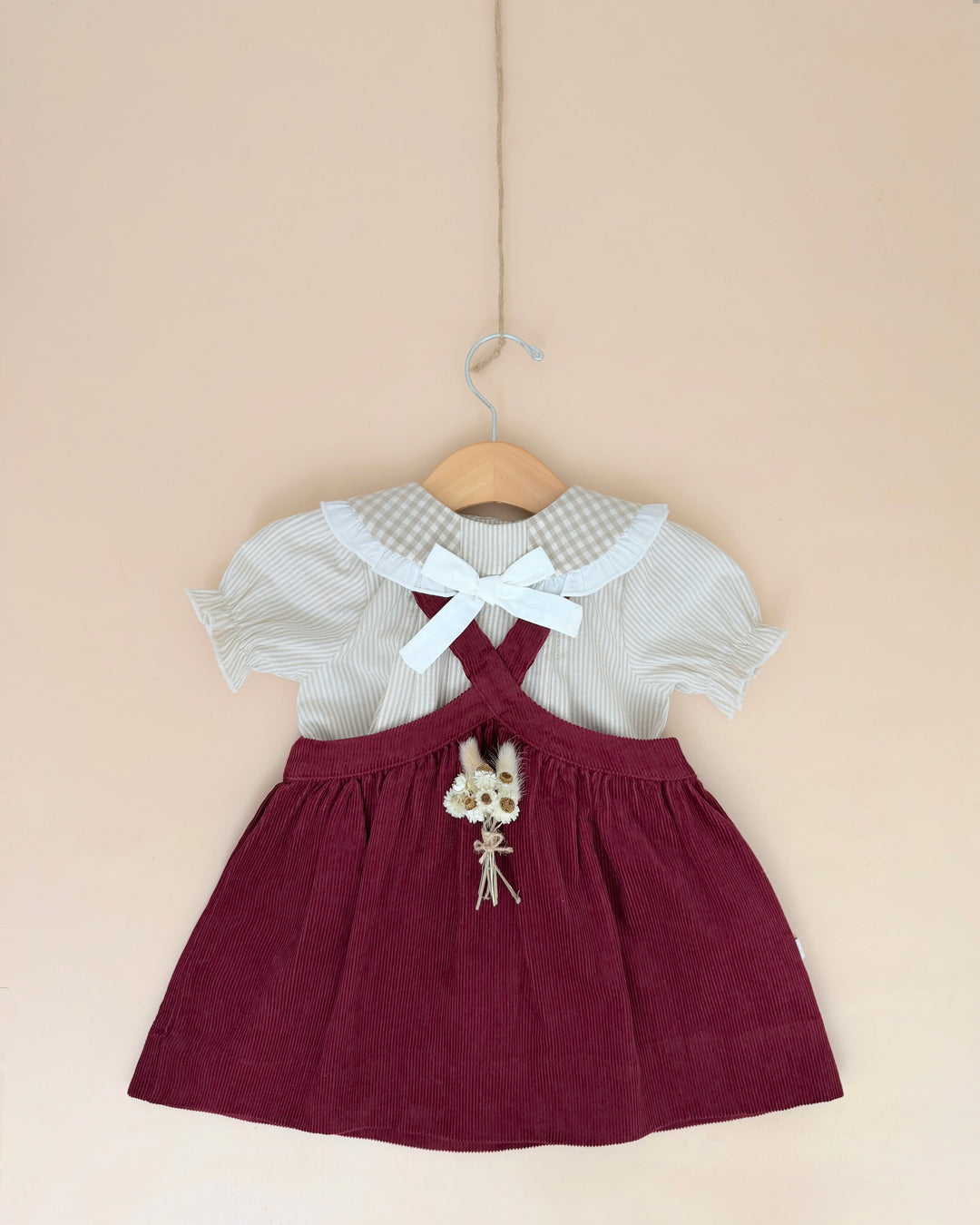 A baby girl's dress hanging on a wooden hanger against a solid beige wall. The dress features a deep burgundy corduroy pinafore skirt and a white and gray gingham short-sleeve top with a white bow at the collar.