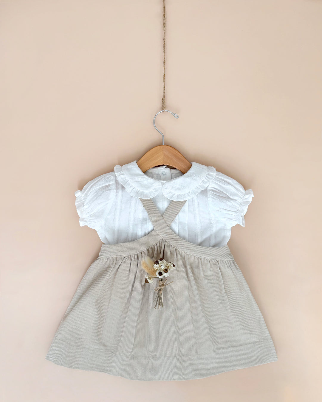 A toddler girl's dress hanging on a wooden hanger against a soft beige wall. The dress features a white short-sleeve top with a ruffled Peter Pan collar and crisscross back straps, paired with a light beige linen-look skirt adorned with a small dried floral accent at the waist.