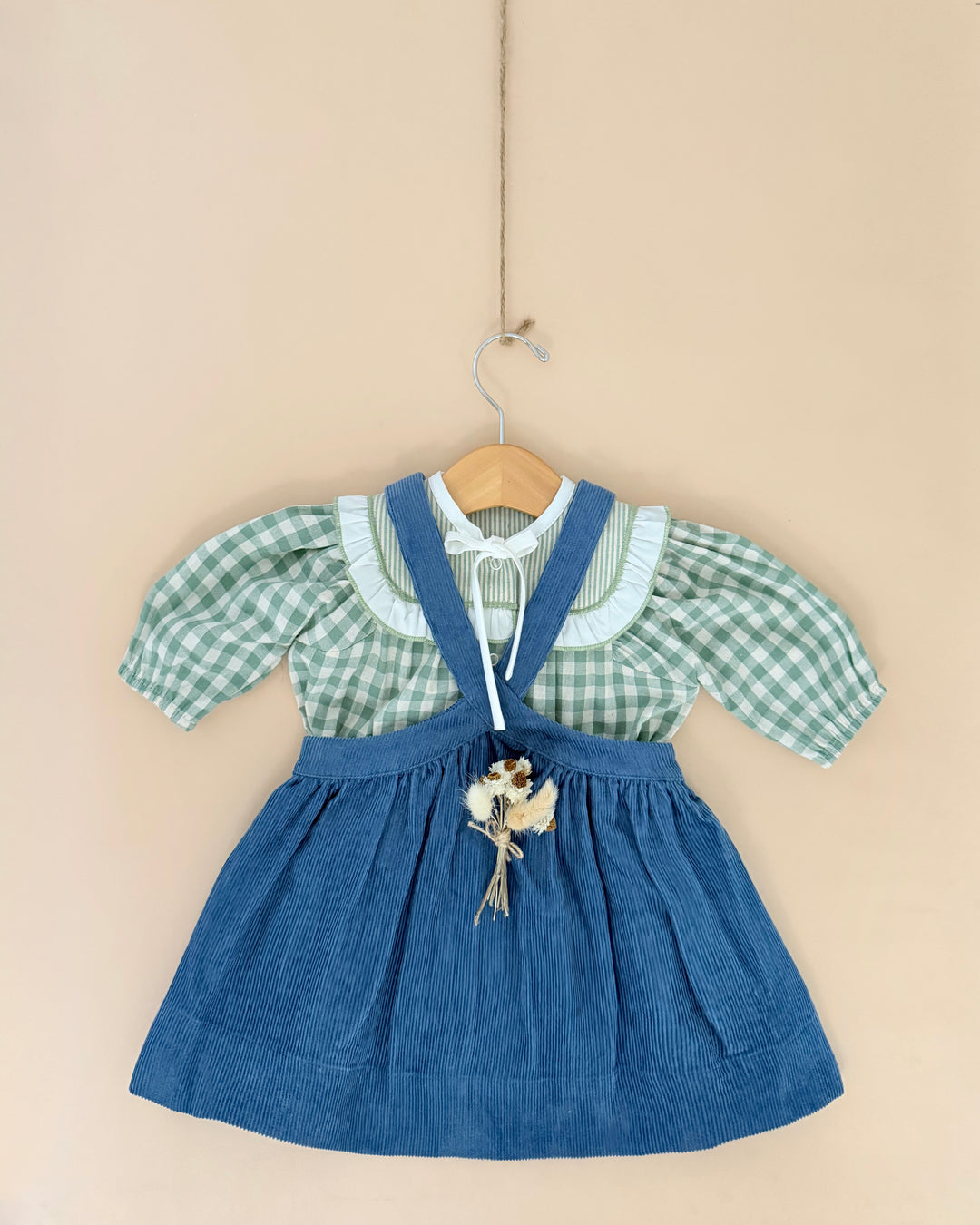 A toddler girl's dress hanging on a wooden hanger against a solid beige wall. The dress features a light green and white gingham short-sleeve blouse layered under a blue denim-style pinafore-style skirt with shoulder straps and a small dried flower accent at the center front.