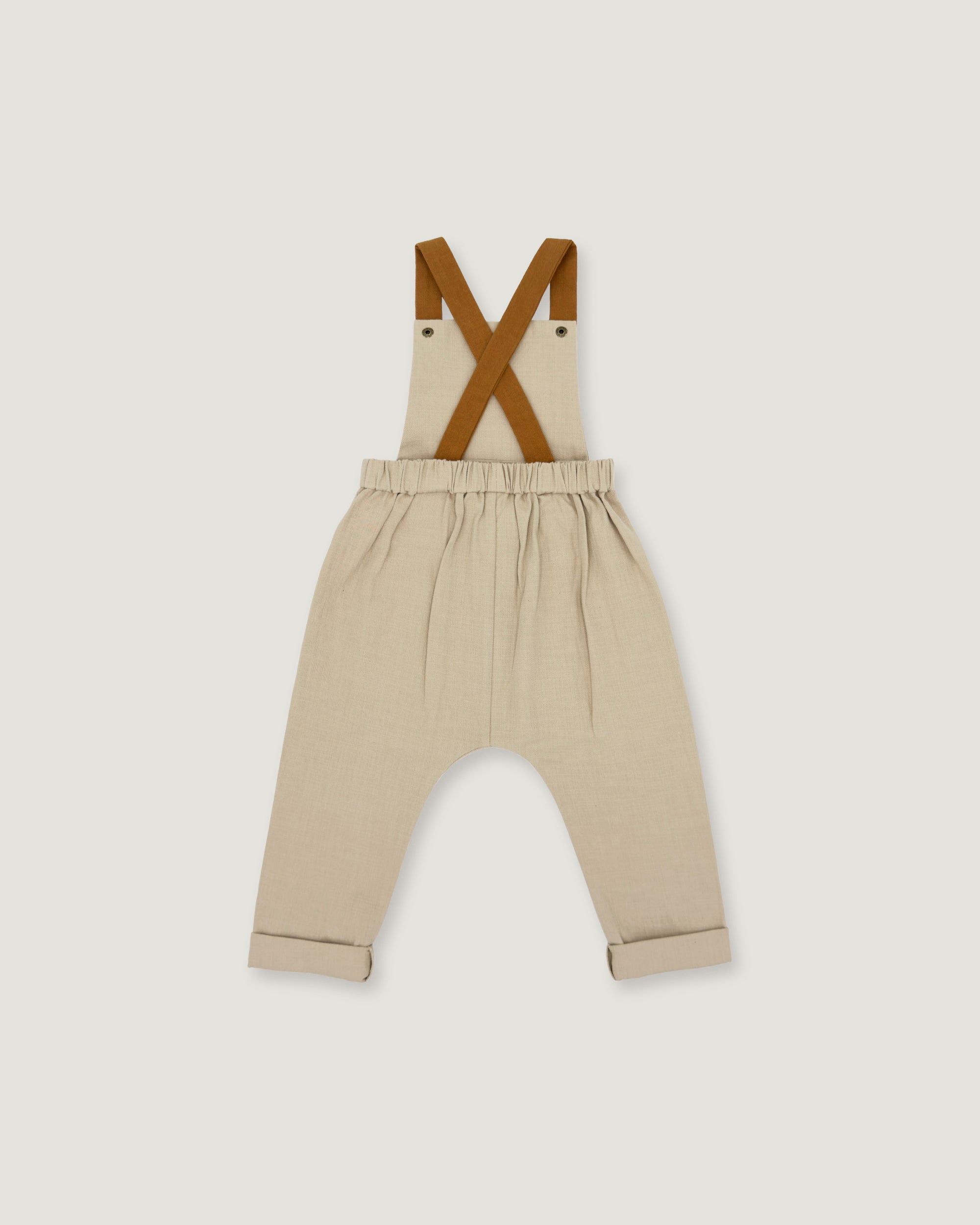 A pair of beige toddler pants with brown suspenders, laid flat against a light gray background.