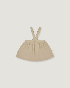 A light beige baby dress with shoulder straps, laid flat against a solid light gray background.