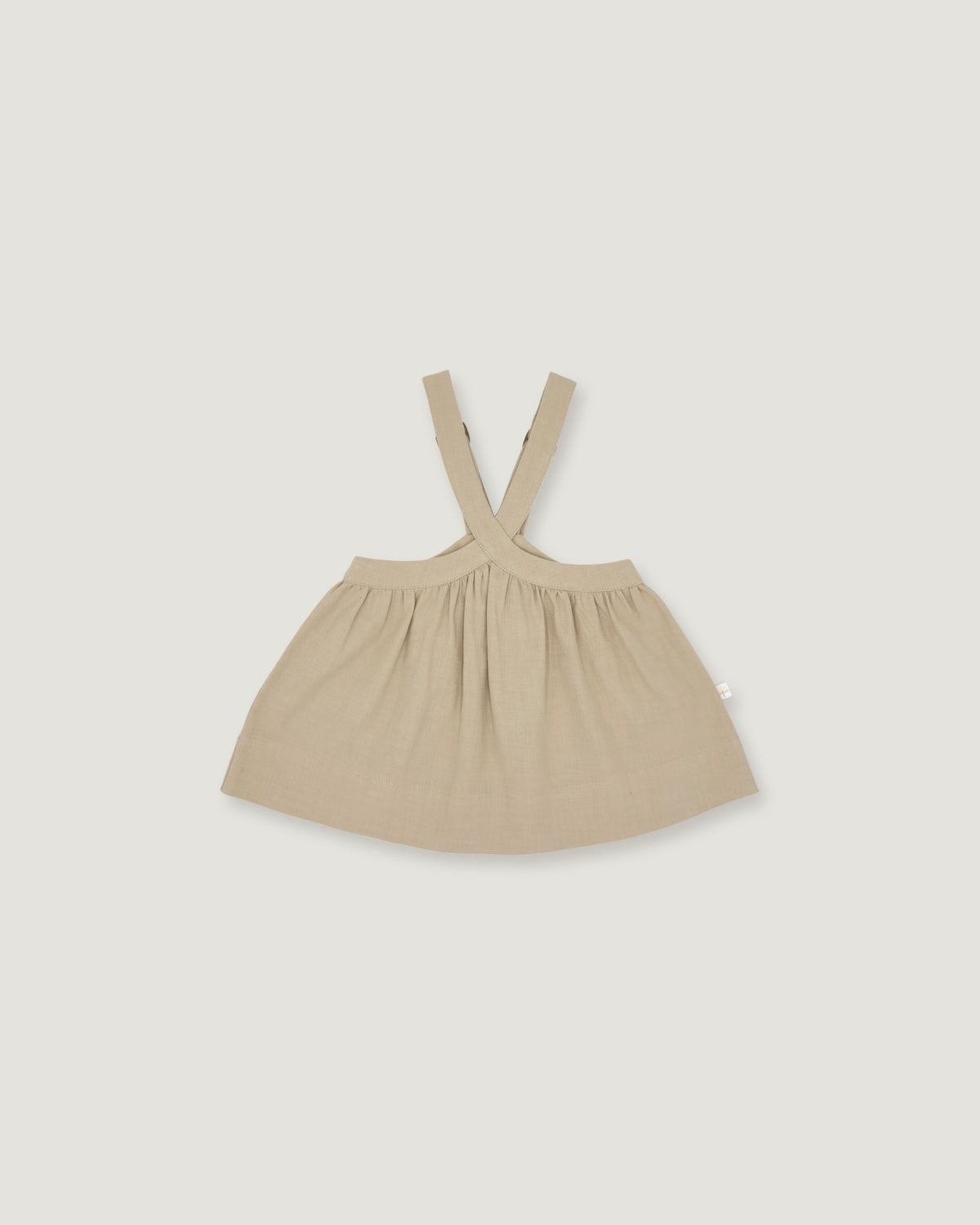 A light beige baby dress with shoulder straps, laid flat against a solid light gray background.