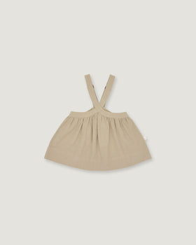 A light beige baby dress with shoulder straps, laid flat against a solid light gray background.