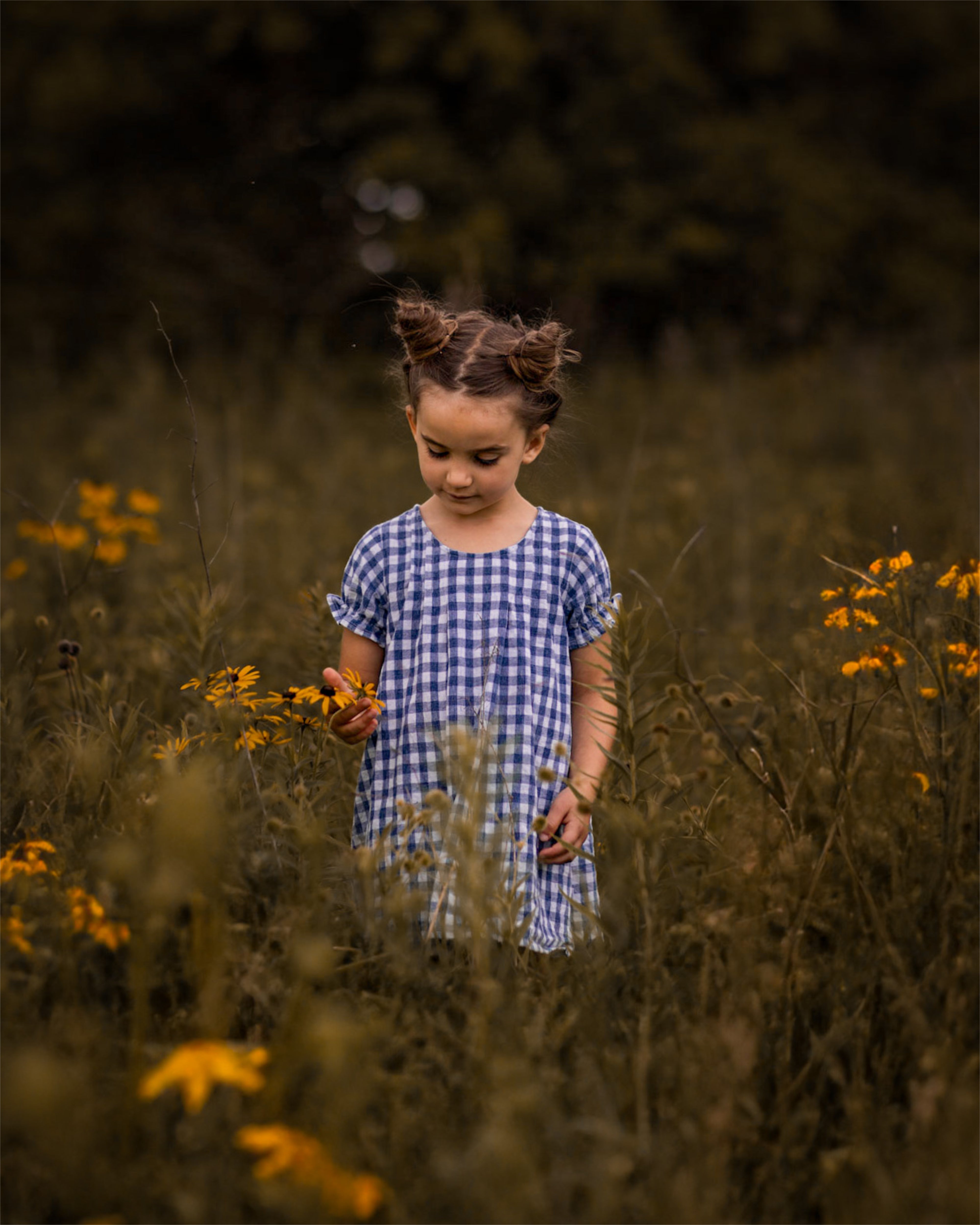 A little girl stands among vibrant flowers, wearing a Blue Gingham linen dress. The back view of the dress reveals its pin-tucked design, a yoke with pleats, and delicate ruffles at the short sleeve hem. Surrounded by colorful blooms, she embodies a joyful spirit, with her hair styled up, enjoying the beauty of nature. The A-line shape of the dress allows for comfort and ease as she plays in this enchanting floral setting.