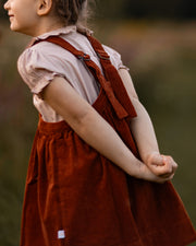 Child wearing a red dress with suspenders against a blurred natural background