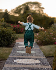 A 3-year-old model boy joyfully jumping to showcase the back of his classic light blue Henley shirt with white Swiss dots, made from natural, sustainable, and high-quality materials. The shirt features a timeless Henley placket, while the spruce corduroy long overalls add a rich texture and warmth. This dynamic pose highlights the outfit's playful style and comfort, perfect for active little ones.