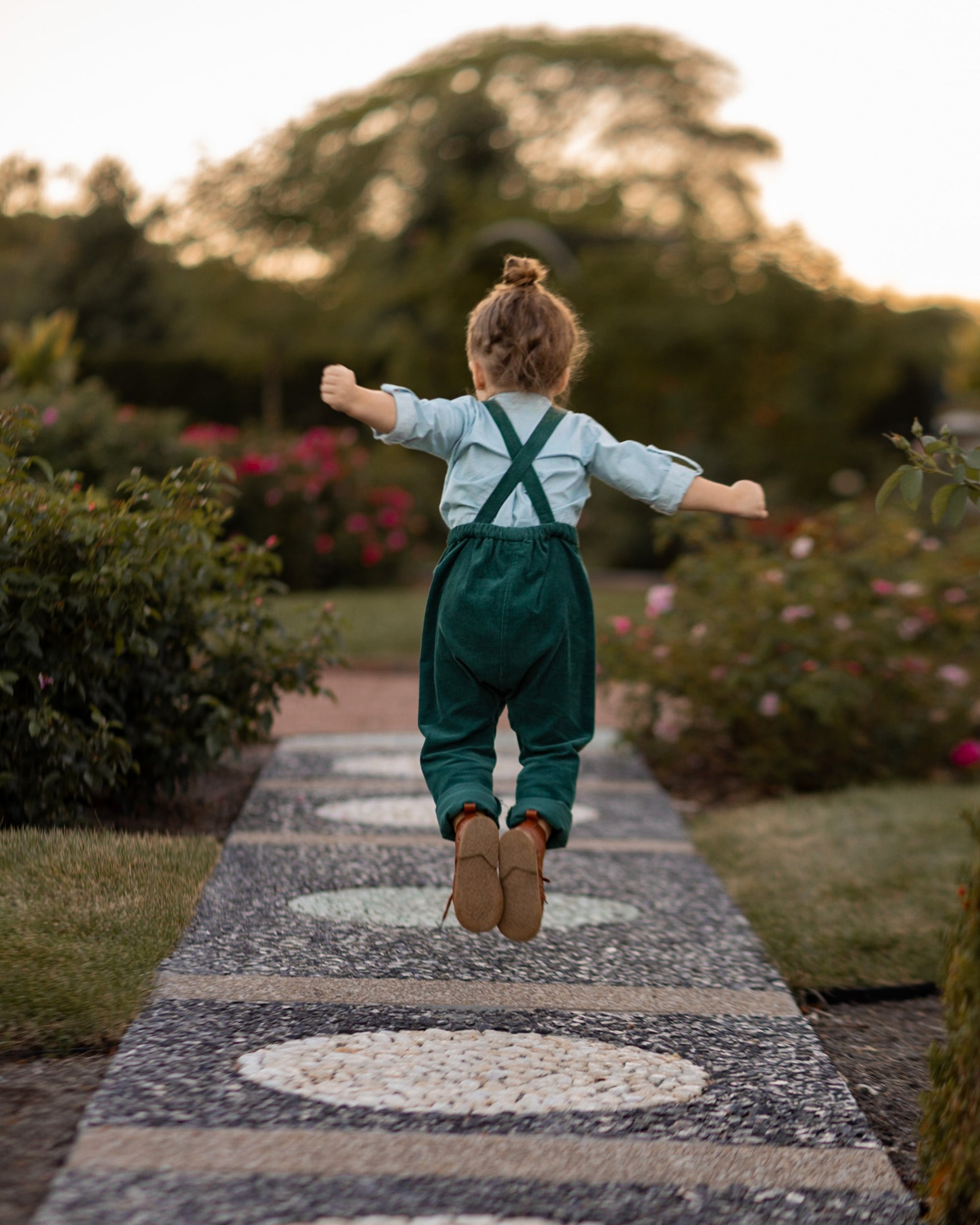 A 3-year-old model boy joyfully jumping to showcase the back of his classic light blue Henley shirt with white Swiss dots, made from natural, sustainable, and high-quality materials. The shirt features a timeless Henley placket, while the spruce corduroy long overalls add a rich texture and warmth. This dynamic pose highlights the outfit's playful style and comfort, perfect for active little ones.