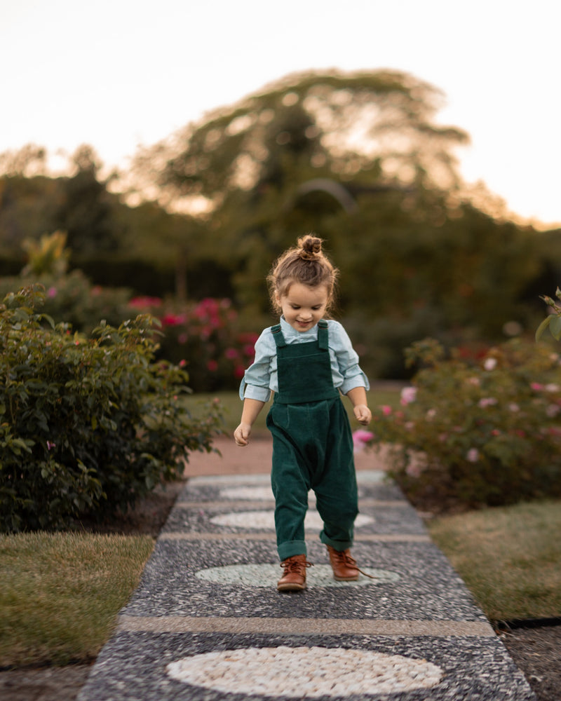 A 3-year-old model boy joyfully jumping outside in a classic light blue Henley shirt with white Swiss dots, made from natural, sustainable, and high-quality materials. He pairs it with spruce corduroy long overalls, which add texture and warmth. The playful action captures the spirit of childhood, showcasing the outfit's comfort and style for outdoor adventures.