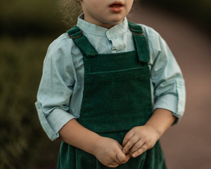 A 3-year-old model boy showcasing a classic light blue Henley shirt with white Swiss dots, crafted from natural, sustainable, and high-quality materials. He pairs it with spruce corduroy long overalls, which add a rich texture and warmth to the outfit. The combination highlights the playful yet stylish nature of children's fashion, perfect for both casual outings and special occasions.