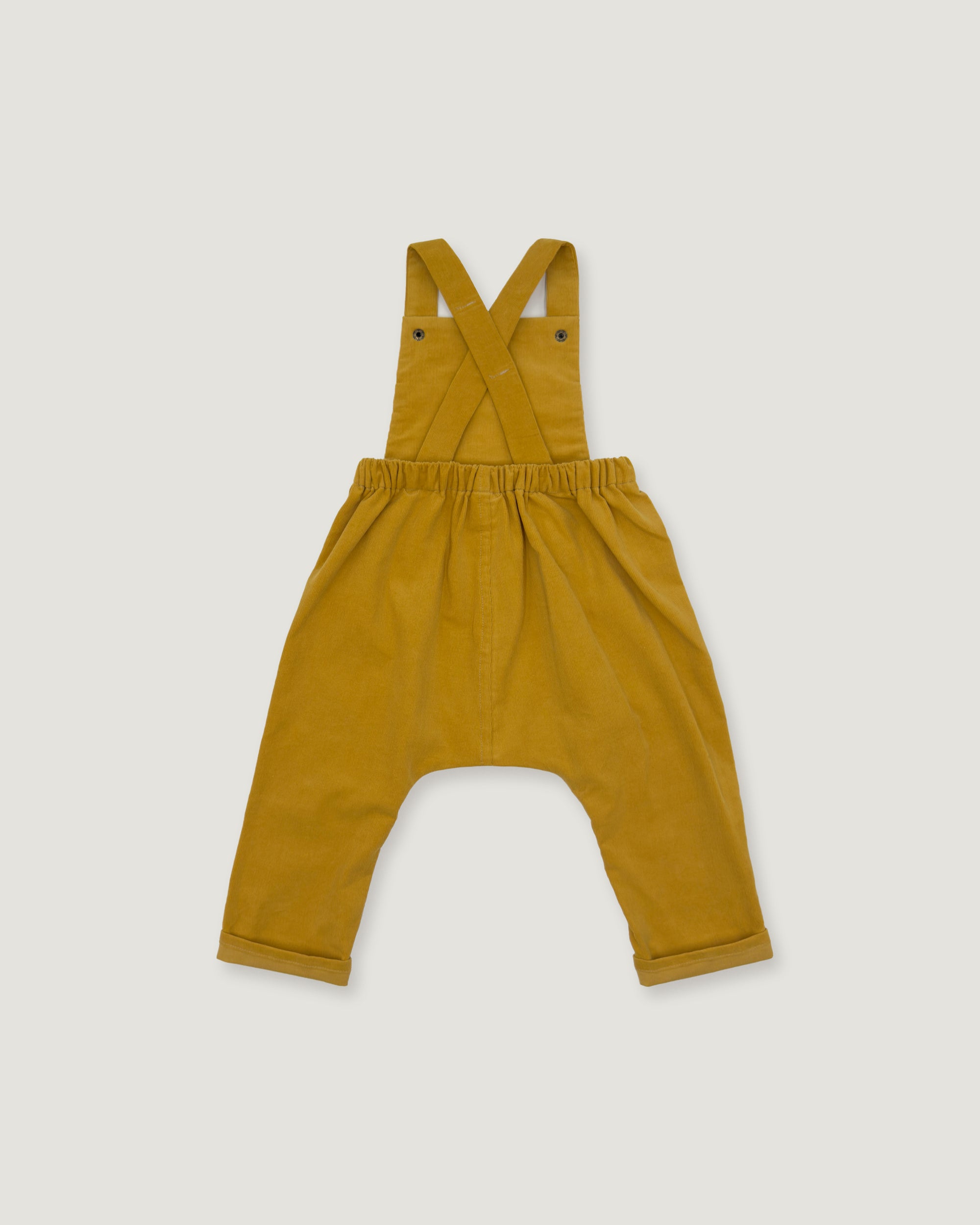 A mustard yellow baby romper with shoulder straps that cross over at the back, an elastic waistband, and wide legs with rolled cuffs, shown flat on a light gray background.