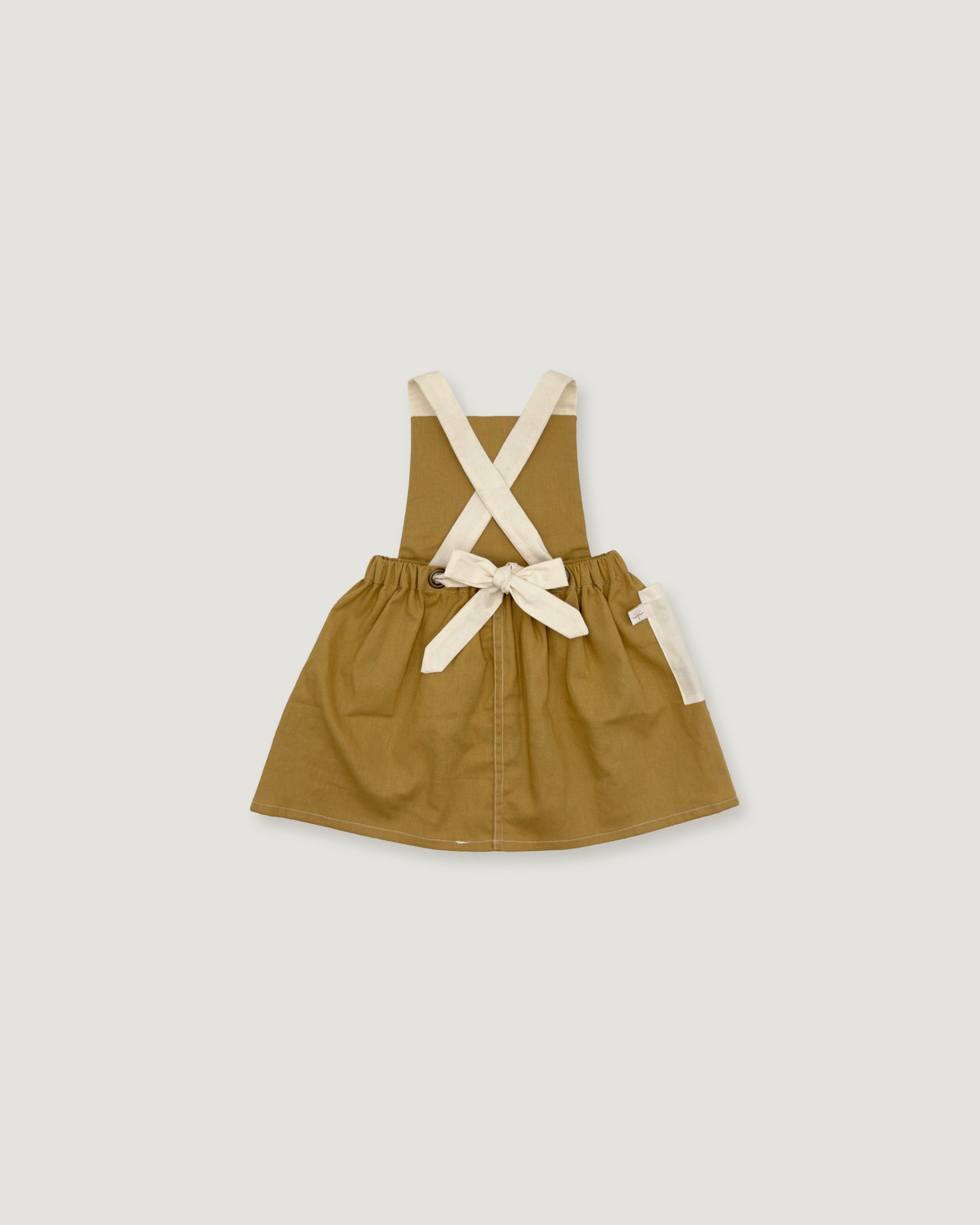 A toddler's utility pinafore dress in olive green with cream-colored shoulder straps and a matching bow at the waist, displayed flat against a light gray background.