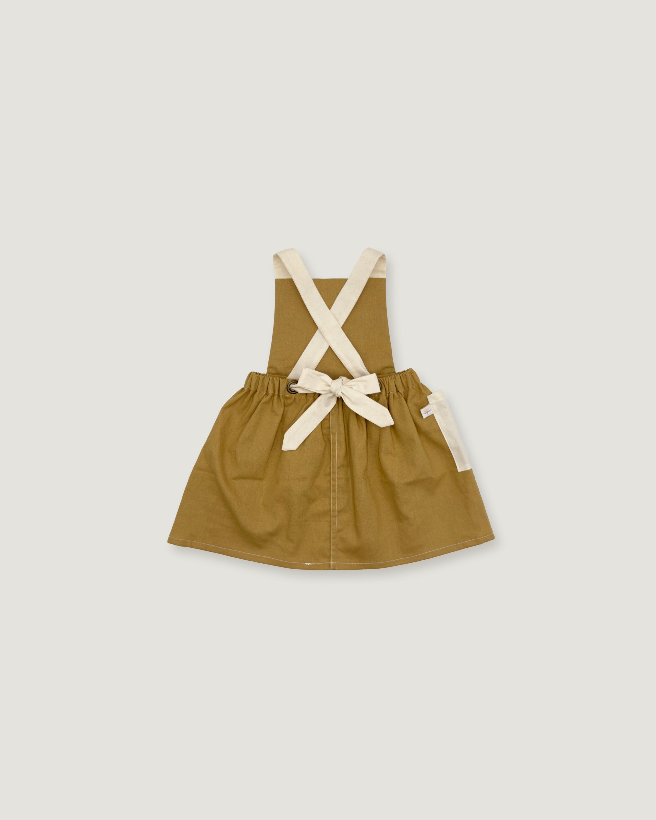 A toddler's utility pinafore dress in olive green with cream-colored shoulder straps and a matching bow at the waist, displayed flat against a light gray background.