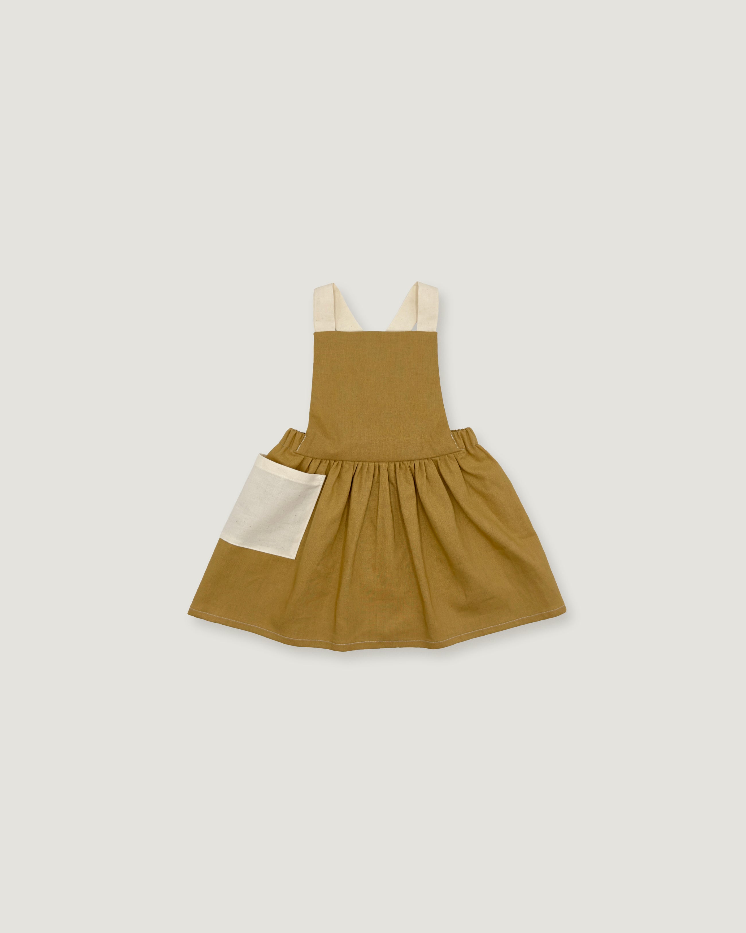 A toddler's sleeveless pinafore dress in a warm mustard-brown color, featuring a gathered skirt, a cream-colored front patch pocket, and cream shoulder straps, shown flat on a light gray background.