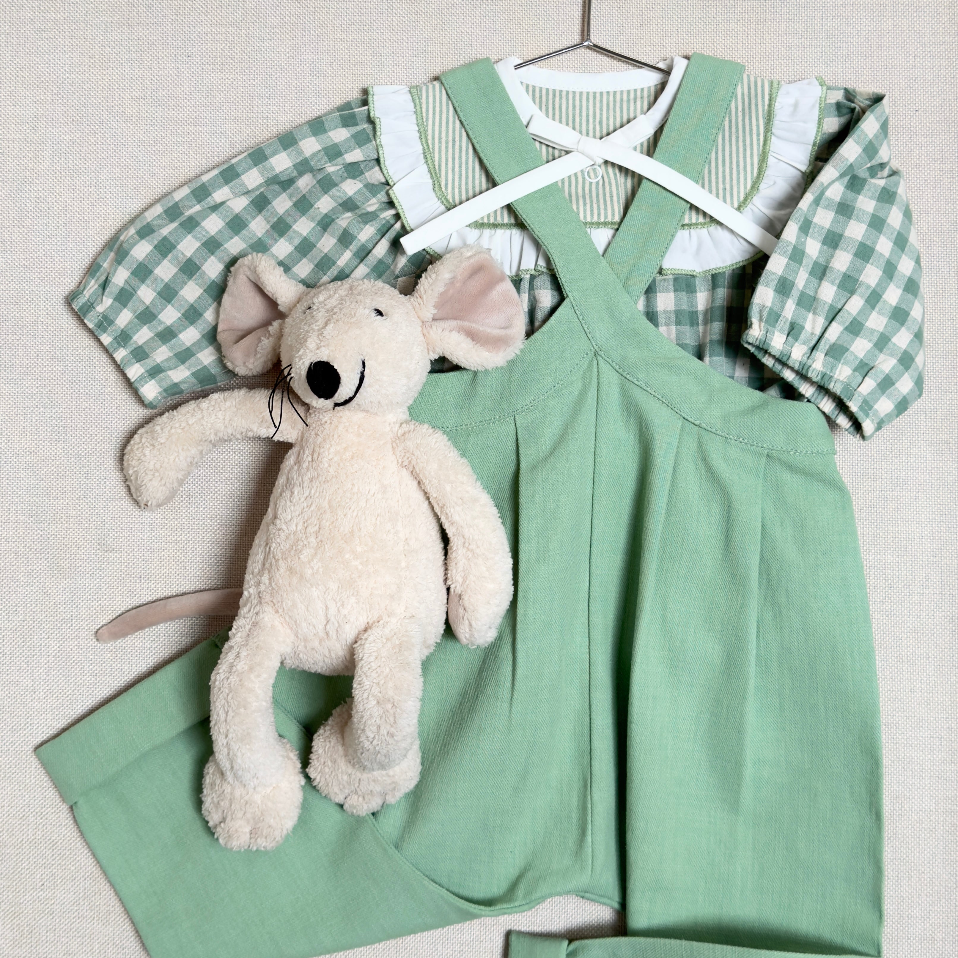 Green checkered shirt and green overalls with a plush toy on a hanger against a light background