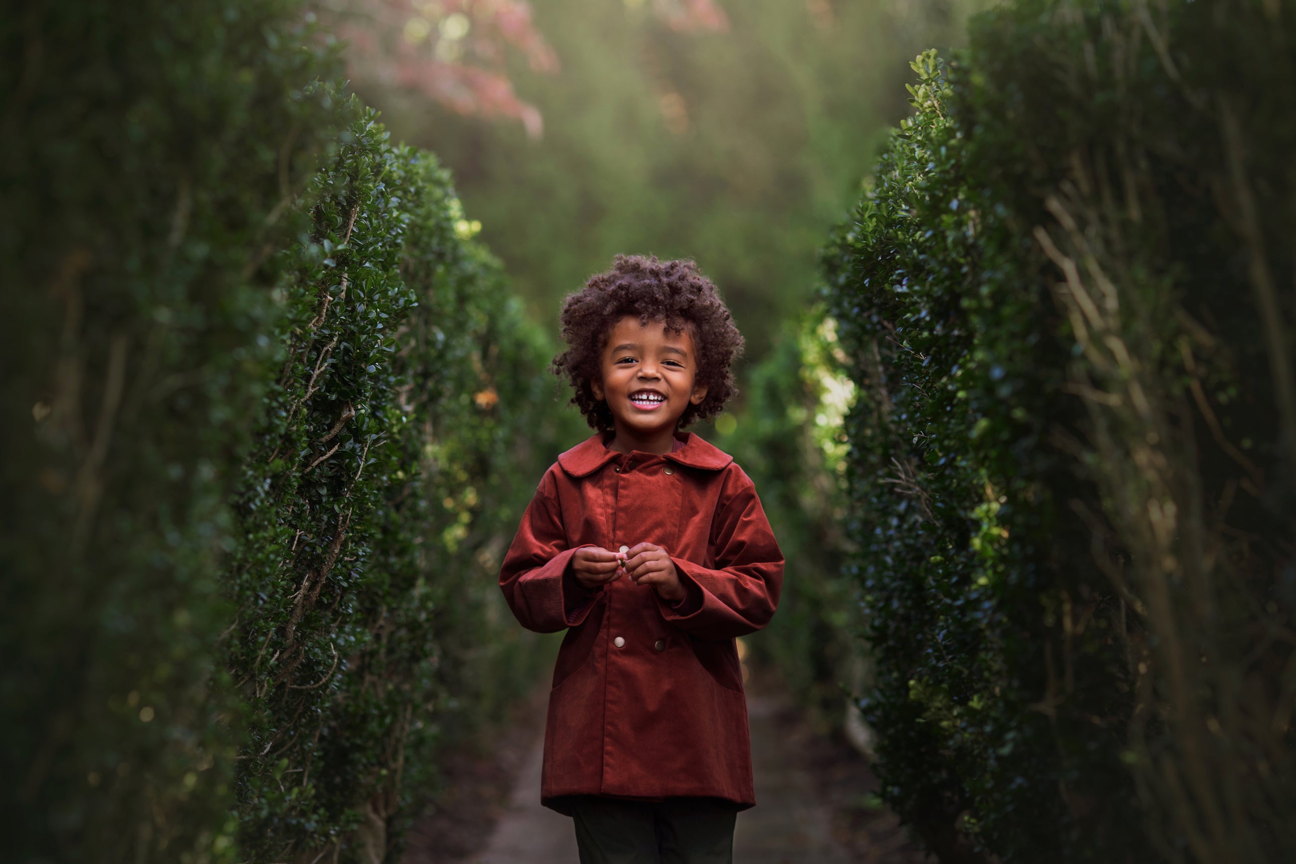 Child in a red coat standing between two tall green bushes