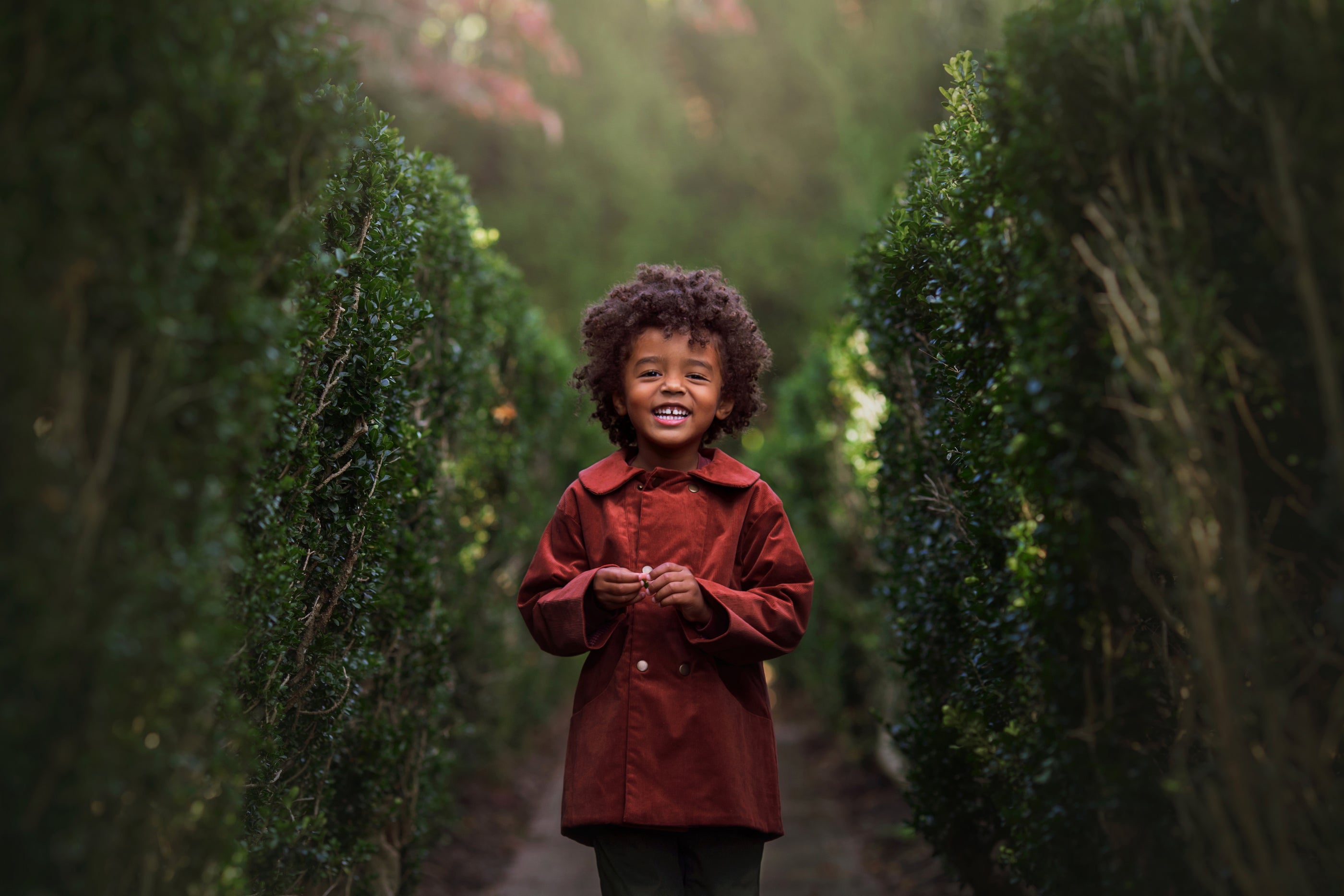 Child in a red coat standing between two tall green bushes
