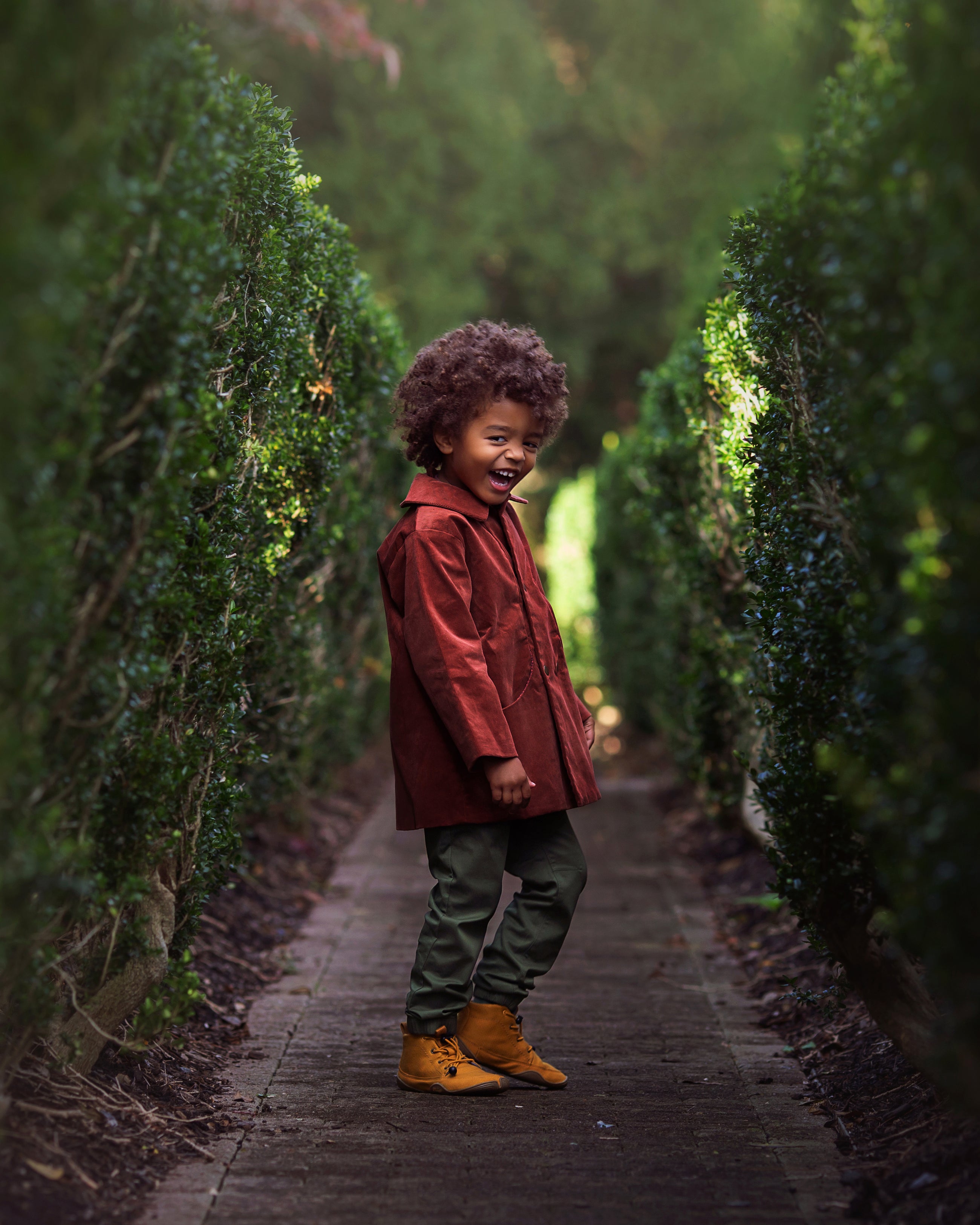 Child in a red coat standing in a garden path lined with green hedges