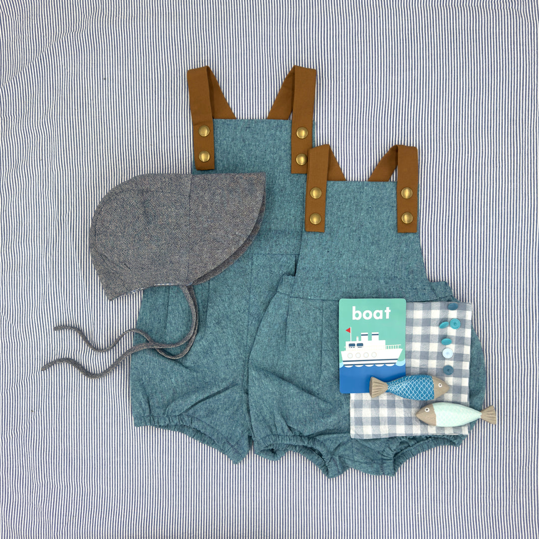 Children's blue romper with brown straps, gray bonnet, and toy book on a striped background