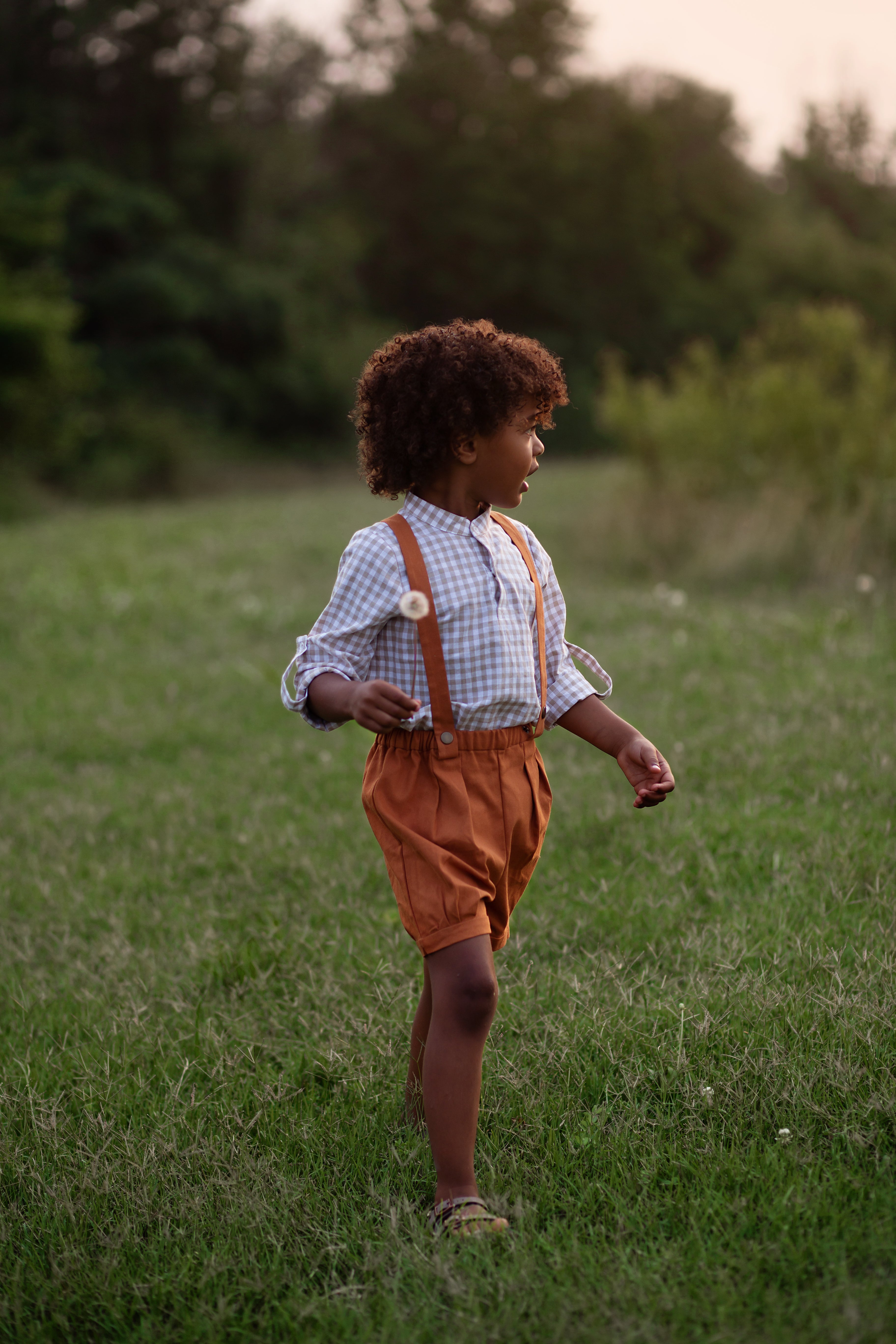 Model boy wearing the Burnt Orange Suspender Shorts with Beige Gingham Henley Shirt, side view - This image captures the side profile, showcasing the adjustable straps and overall silhouette of the outfit