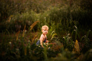 Close-up of a little boy's sweet face in a cornfield wearing chambray blue overalls - Handmade from repurposed linen fabric, these gender-neutral overalls are perfect for passing down. The breathable fabric ensures comfort while exploring the outdoors. Stylish pleats add elegance, and the durable leather-looking straps enhance visual appeal. Adjustable straps in two lengths provide a great fit, making these overalls a versatile and sustainable choice for active kids enjoying summer adventures.
